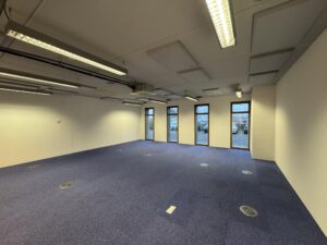 Empty office space with blue carpet, white walls, ceiling lighting, and windows facing De Ruijterkade.
